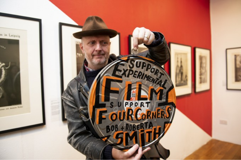 Photograph of the artist Bob and Roberta Smith holding a film canister with the words 'Support experimental film, support Four Corners' written on it. 
