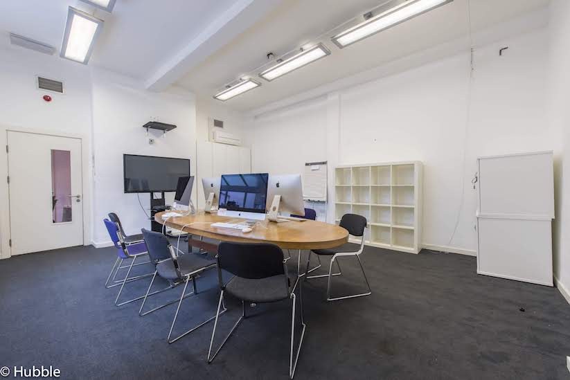 Four Corners' meeting room with white walls, grey carpet, a large wooden table in the centre with computers on top of it, surrounded by grey chairs. There is a large tv screen by one wall. , and a white bookcase by another. It is well-lit and airy.