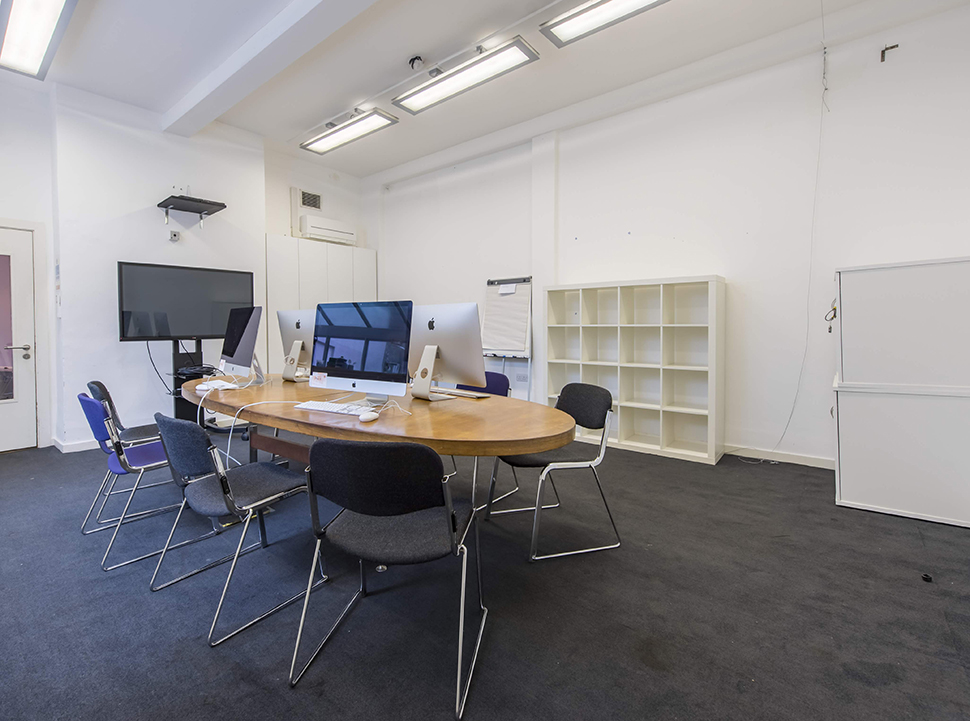 Meeting room with large round table surrounded by chairs. Bookshelf on one wall, TV monitor by another.