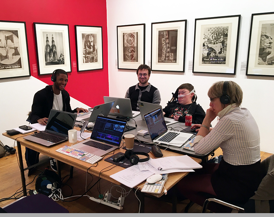 Four smiling people, seated in front of computers. Two of the people are wearing headphones..