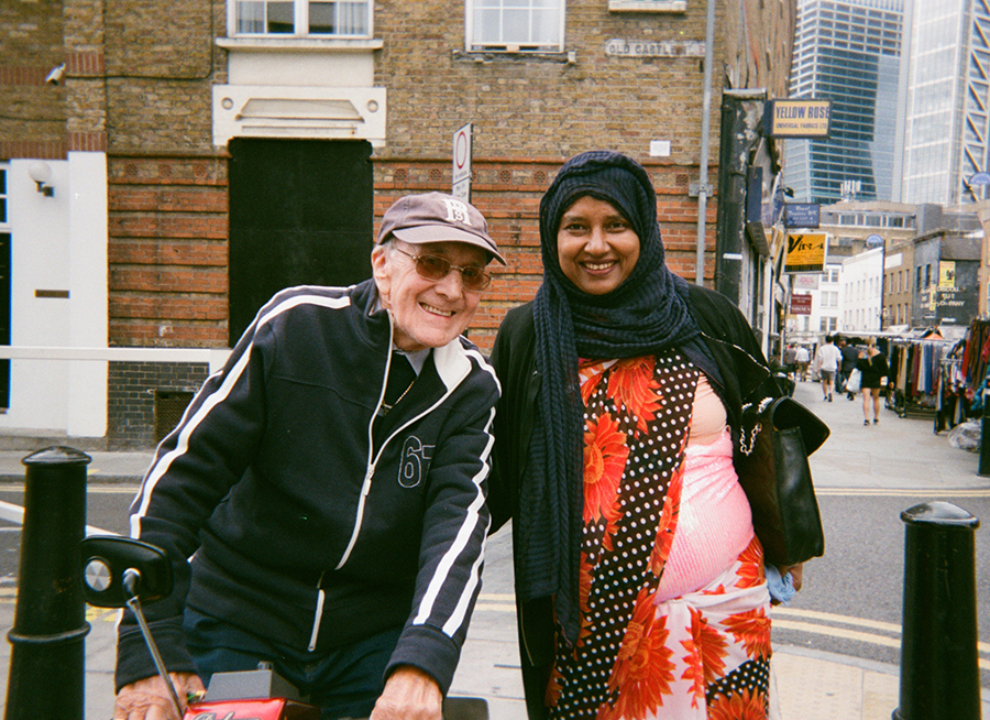 A woman in a headscarf and an older man on a mobility scooter stand next to each other smiling. They are standing in a street and there is a market in the background.  