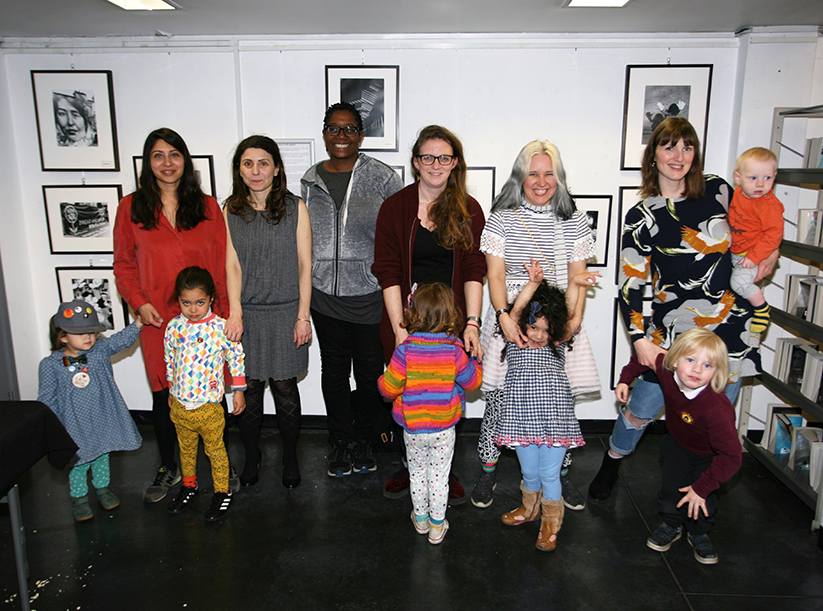 Six mums and six small children stand in front of a wall displaying framed black and white photographs. 