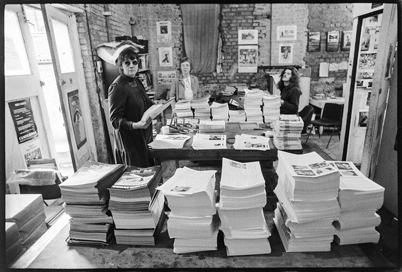 Black and white archive photograph of Jo Spence, Shirley Read and Ed Barber, standing behind tall stacks of Camerawork magazine.