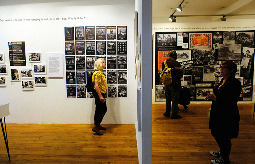 Three people standing in a gallery looking at a collage of posters.