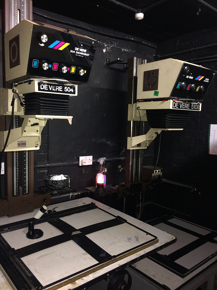 Darkroom stop, dev, and fix trays in use, with three enlargers against the back wall.