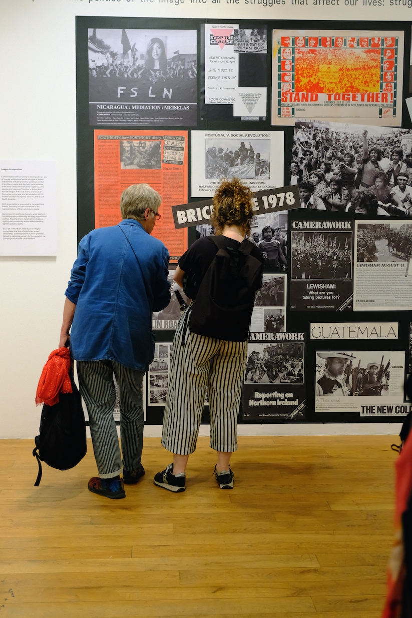 Two people facing away from the camera, looking at a large collage of posters on the gallery wall. 