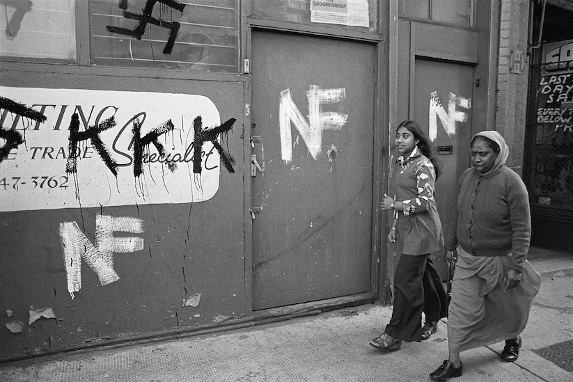 Black and white photograph of two women of colour walking down a street in East London. 'KKK,' 'NF,' and a swastika are painted on the wall.