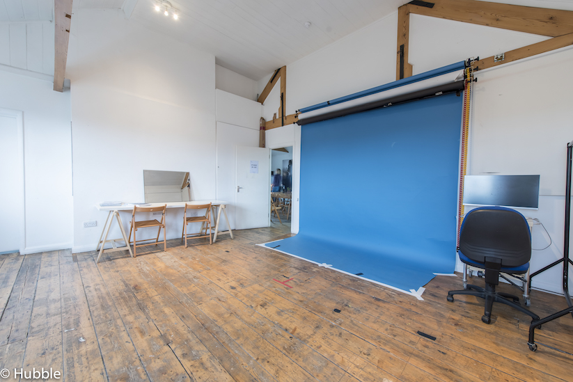 Photography studio with white walls, wooden floors, and high ceilings. There is a blue backdrop on the right hand wall, and a desk with a mirror on the left.